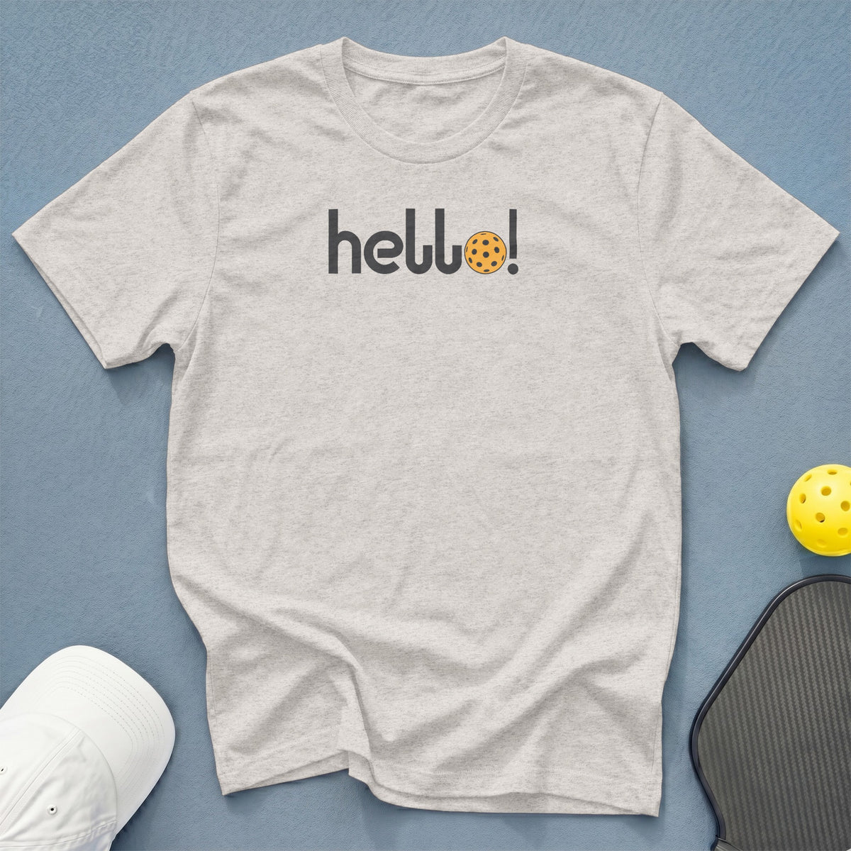 Light grey t-shirt with "hello!" and pickleball graphic, next to a pickleball and paddle.