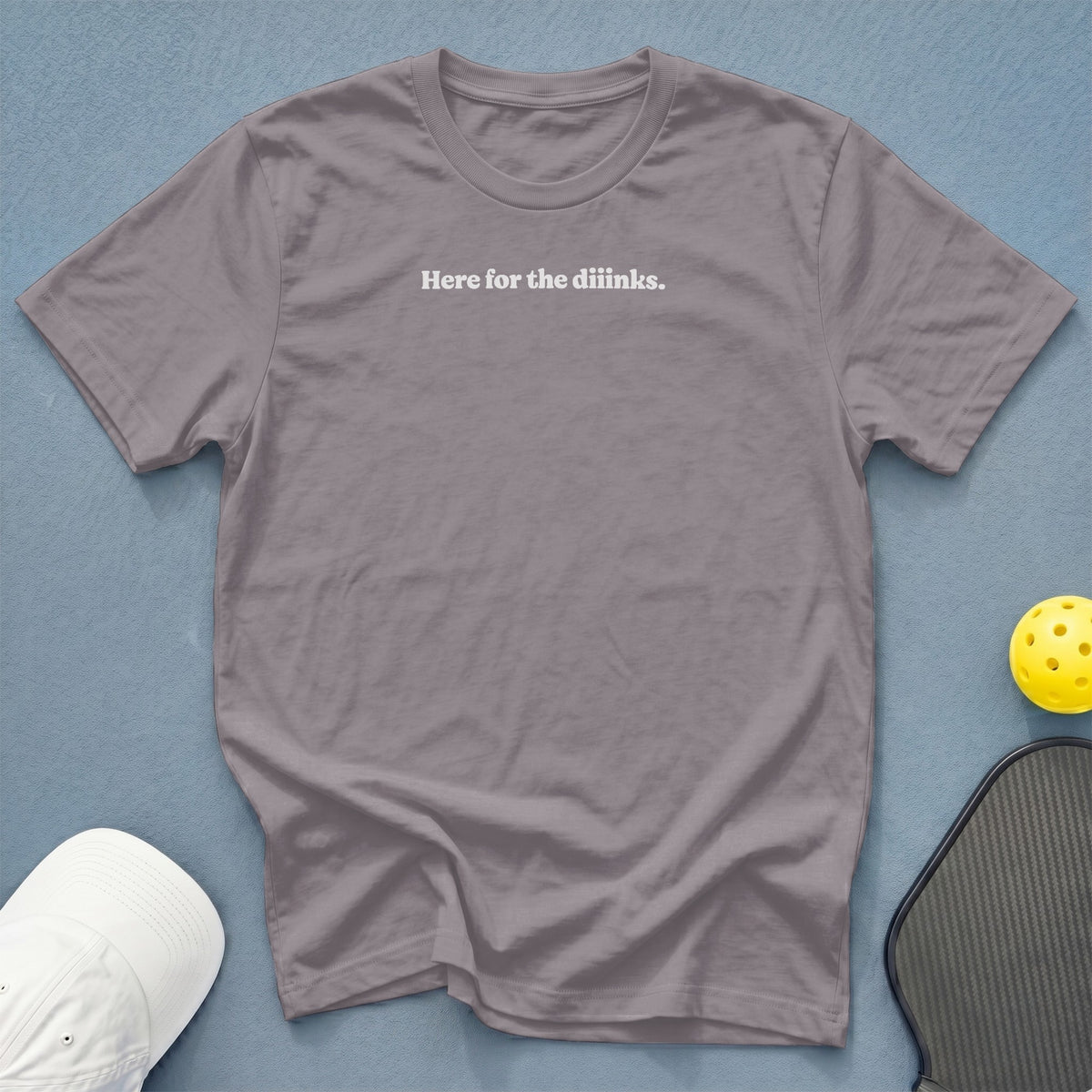 Gray t-shirt with white text "Here for the diinks" near pickleball gear.