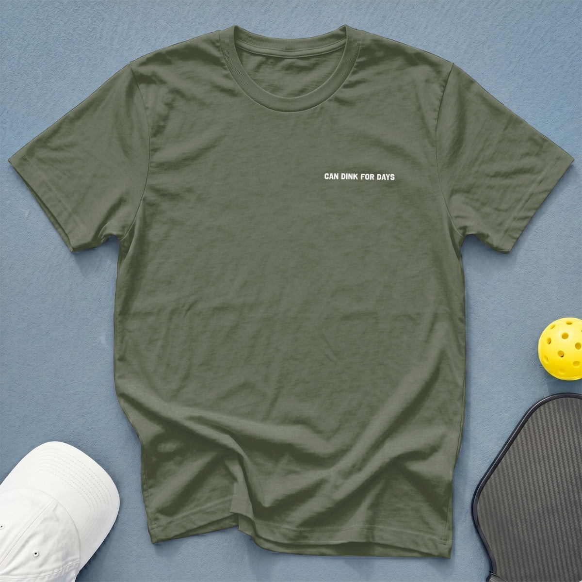 Olive green t-shirt with "CAN DINK FOR DAYS" text, next to a pickleball and paddle.