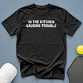 Black t-shirt with white text reading "IN THE KITCHEN CAUSING TROUBLE".