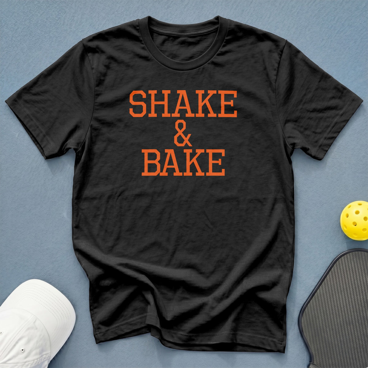Black t-shirt with orange "SHAKE & BAKE" text and pickleball equipment.