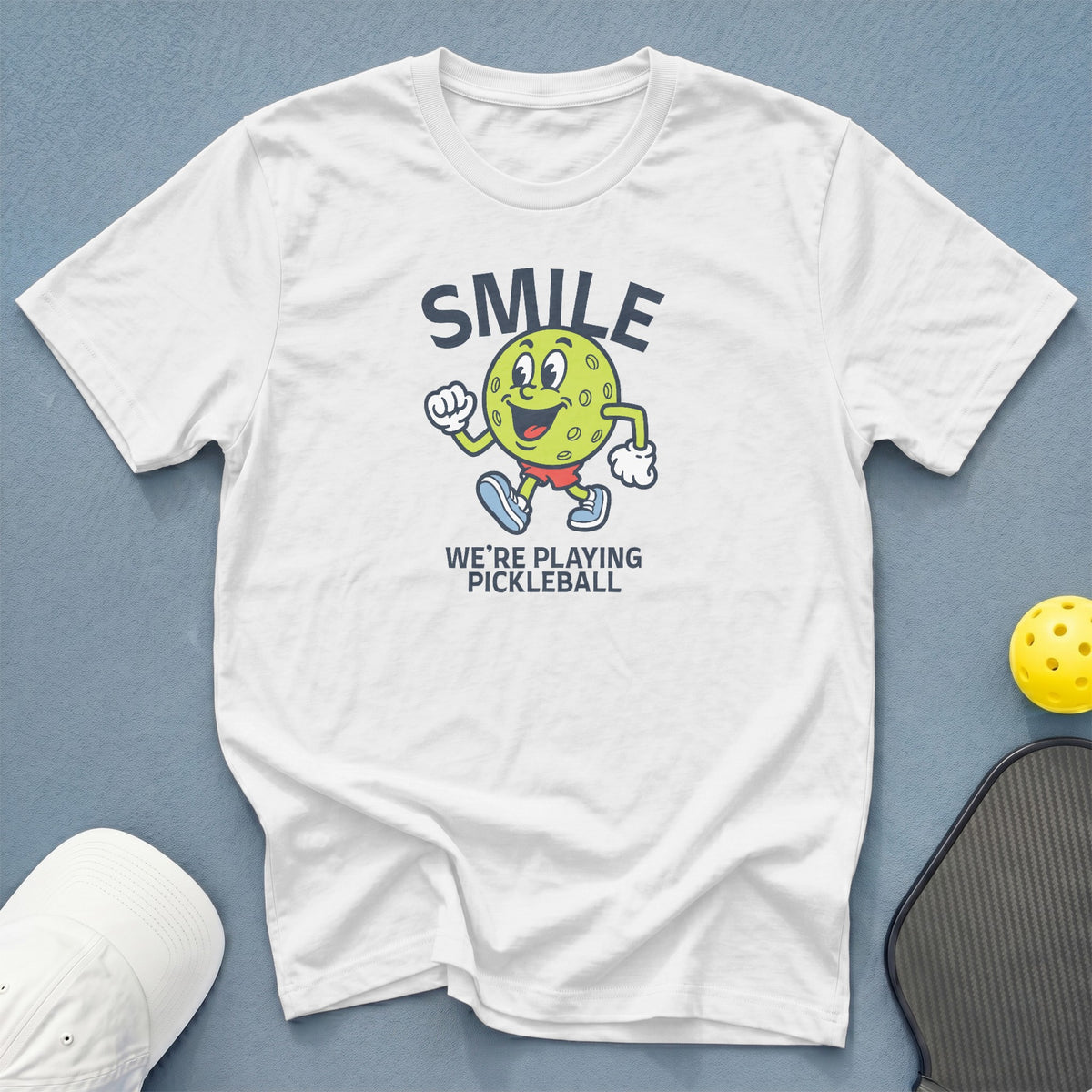 White t-shirt with cartoon pickleball character saying "SMILE WE'RE PLAYING PICKLEBALL".