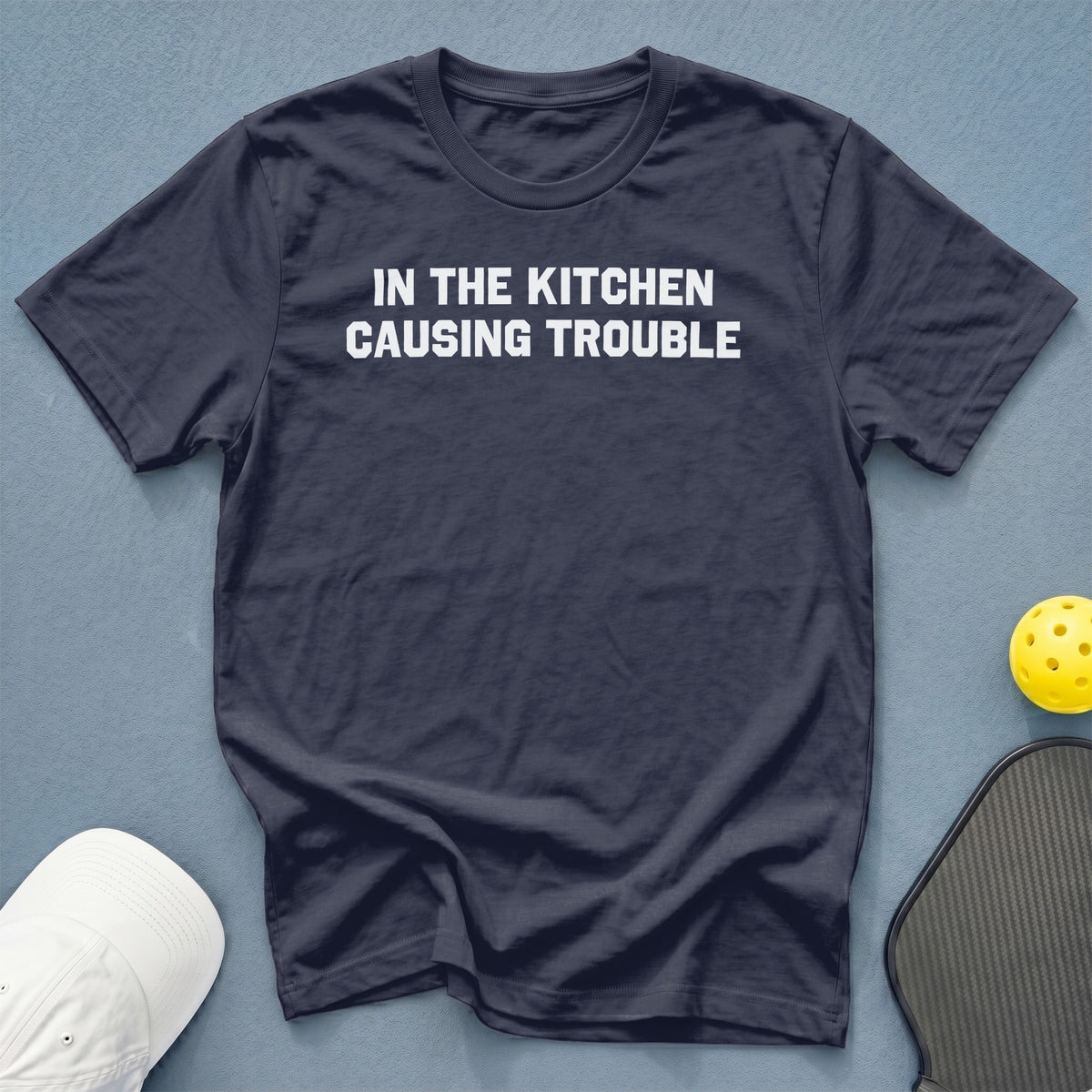 Dark t-shirt with white text reading "IN THE KITCHEN CAUSING TROUBLE."