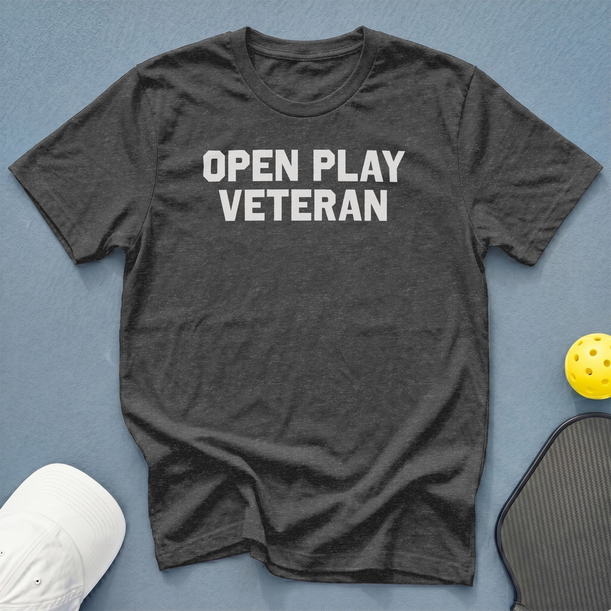 Dark grey t-shirt with "OPEN PLAY VETERAN" text, pickleball paddle, ball, and cap.