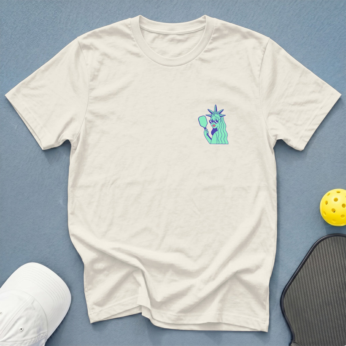 Cream t-shirt with cartoon Statue of Liberty holding pickleball paddle.