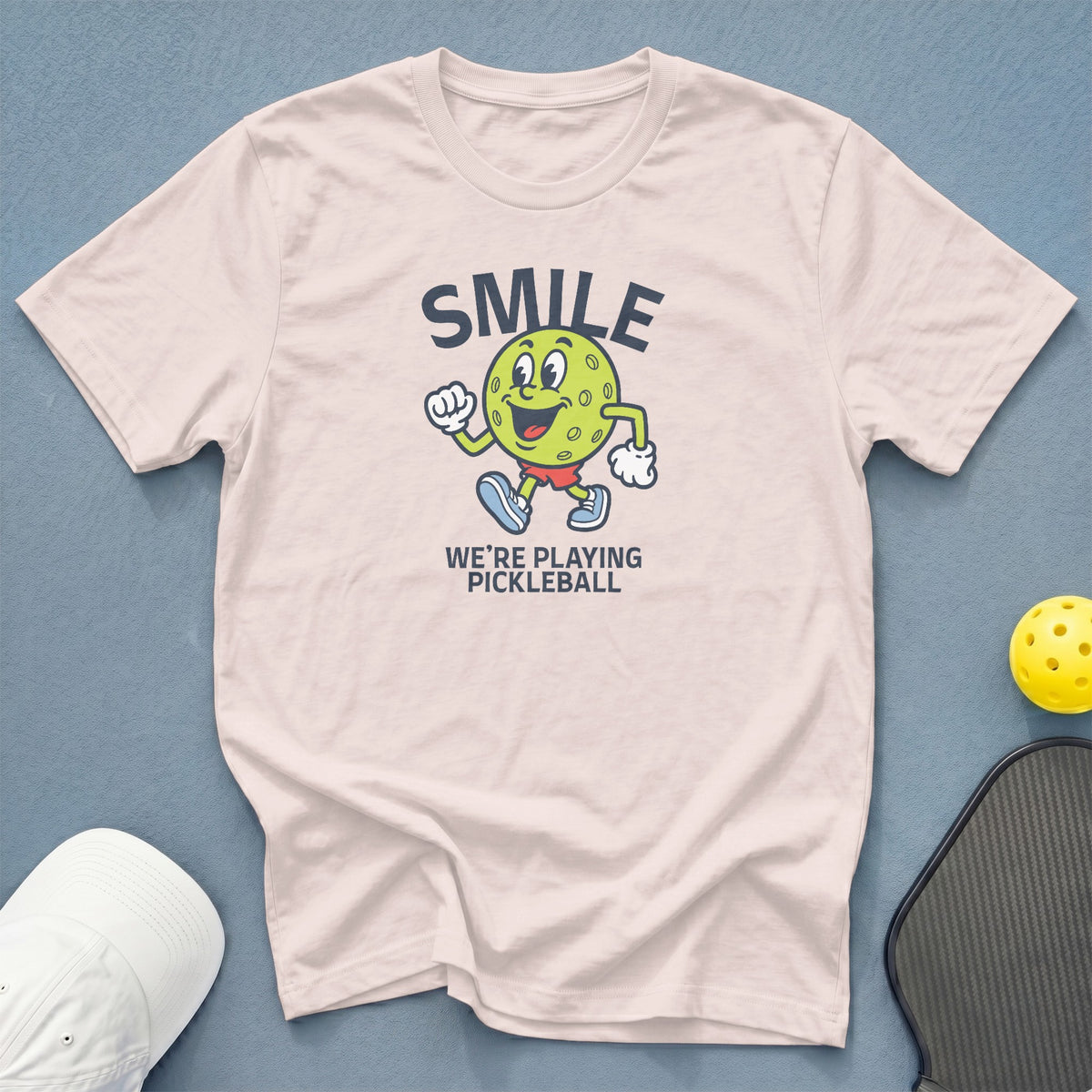 T-shirt with a cartoon pickleball character saying "SMILE WE'RE PLAYING PICKLEBALL".