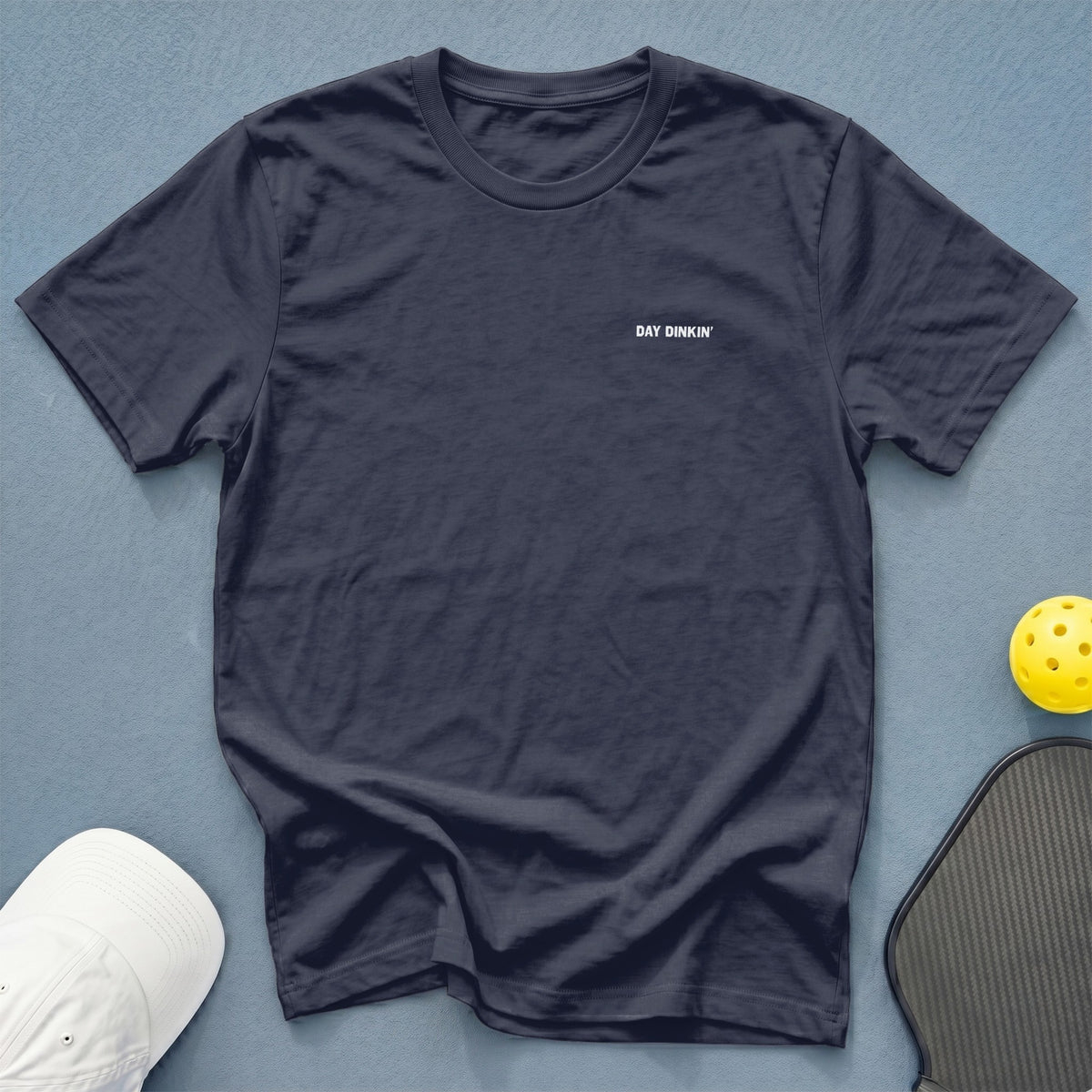 Dark blue t-shirt with "DAY DINKIN'" text, white hat, and pickleball equipment.