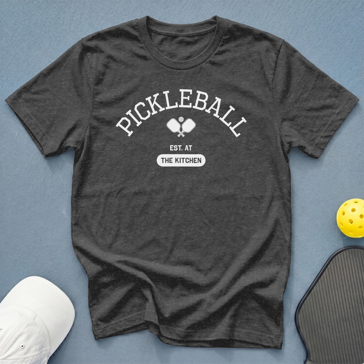 Dark grey t-shirt with "Pickleball Est. At The Kitchen" graphic and pickleball paddles.