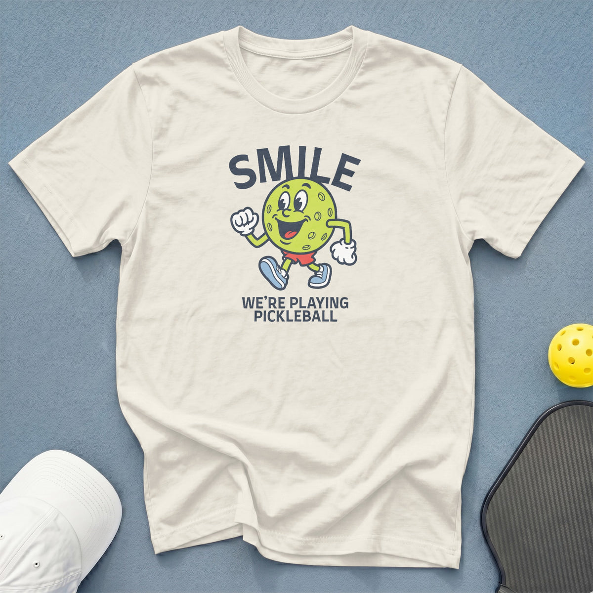 Cream t-shirt with "SMILE WE'RE PLAYING PICKLEBALL" and cartoon pickleball character.