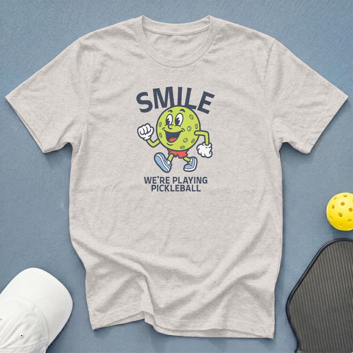 Light gray t-shirt with a graphic of a smiling pickleball and text "SMILE WE'RE PLAYING PICKLEBALL".