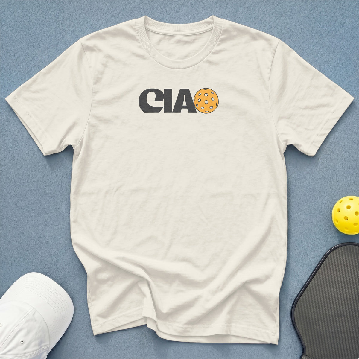 Cream t-shirt with "CIAO" and a pickleball graphic, next to a pickleball and paddle.