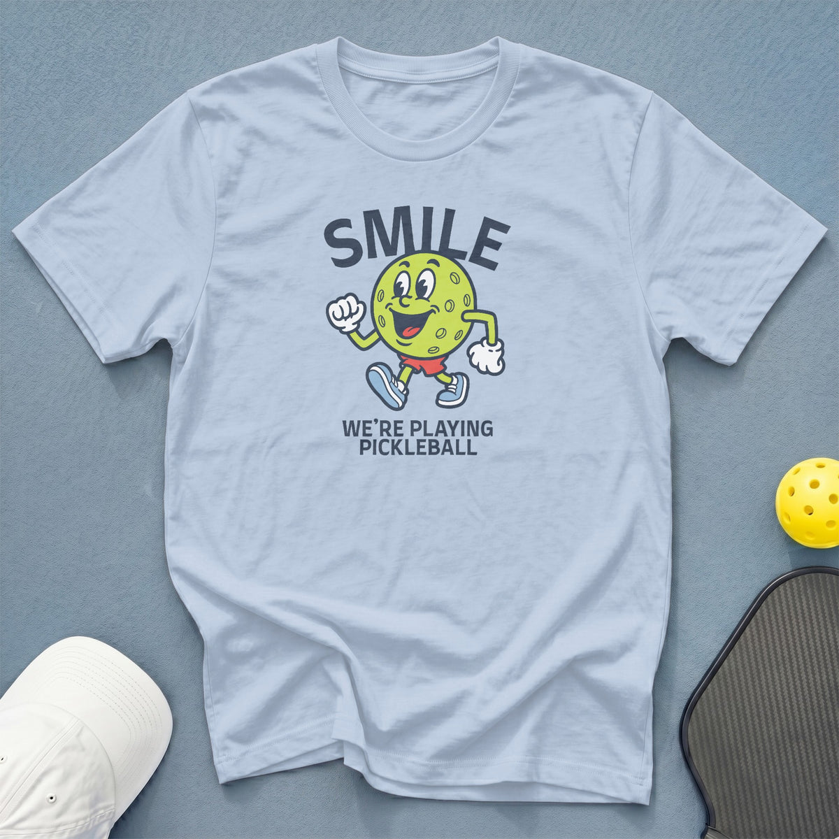 Light blue t-shirt with smiling pickleball character and "SMILE WE'RE PLAYING PICKLEBALL" text.