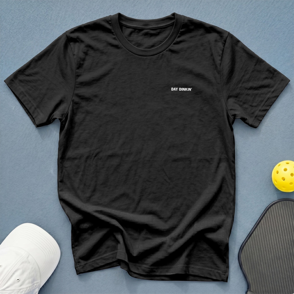 Black t-shirt with white text "DAY DINKIN'" next to pickleball gear.