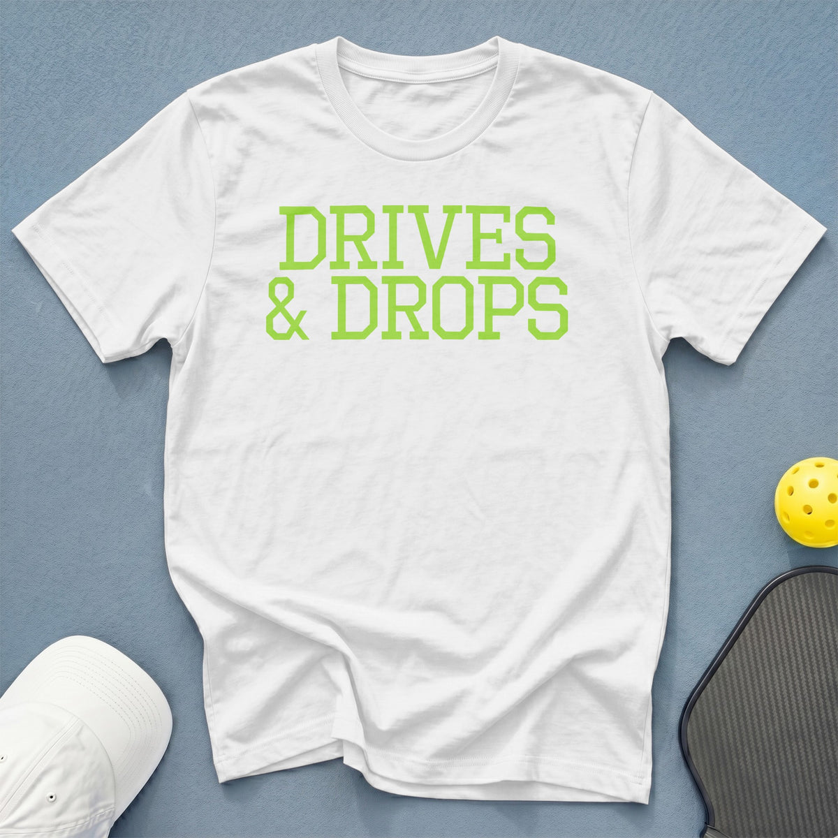 White t-shirt with lime green text "DRIVES & DROPS" and pickleball equipment.